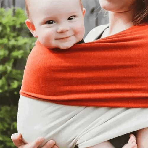 Bébé souriant porté en toute sécurité avec une écharpe de portage rouge et beige, garantissant confort et liberté aux parents.
