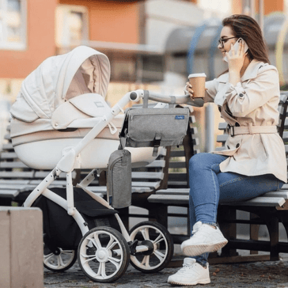 Femme assise près d'une poussette équipée d'un matelas à langer nomade PerfectMat™ gris, tenant une tasse de café et un téléphone.