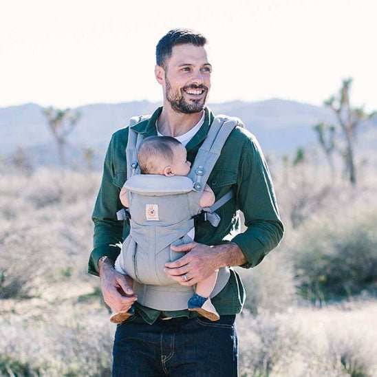 père portant bébé dans un porte-bébé ergonomique gris en plein air avec paysage désertique en arrière-plan