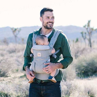 père portant bébé dans un porte-bébé ergonomique gris en plein air avec paysage désertique en arrière-plan