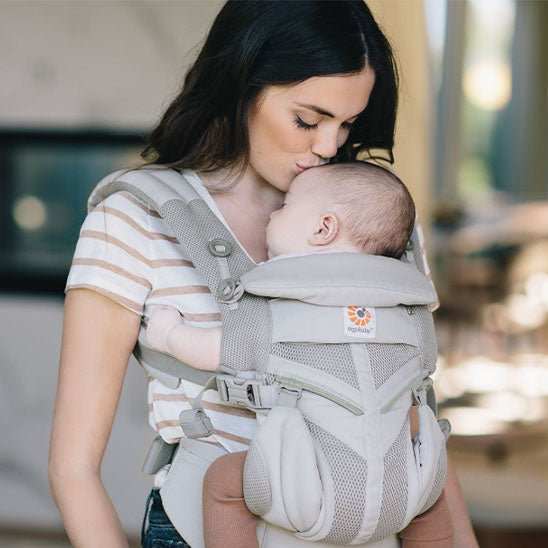 Femme portant un bébé dans un porte-bébé ergonomique gris avec sangles rembourrées et ceinture ajustable, dans un intérieur lumineux.