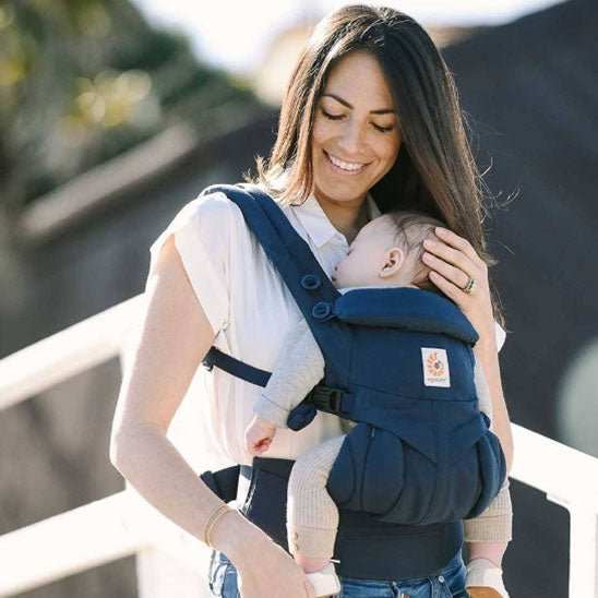 femme souriante portant un bébé endormi dans un porte-bébé ergonomique bleu en extérieur