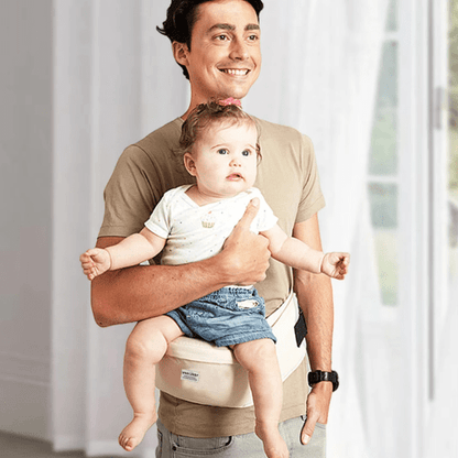 Homme portant un bébé dans un porte-bébé avec tabouret de taille beige, confort ergonomique et soutien optimal.